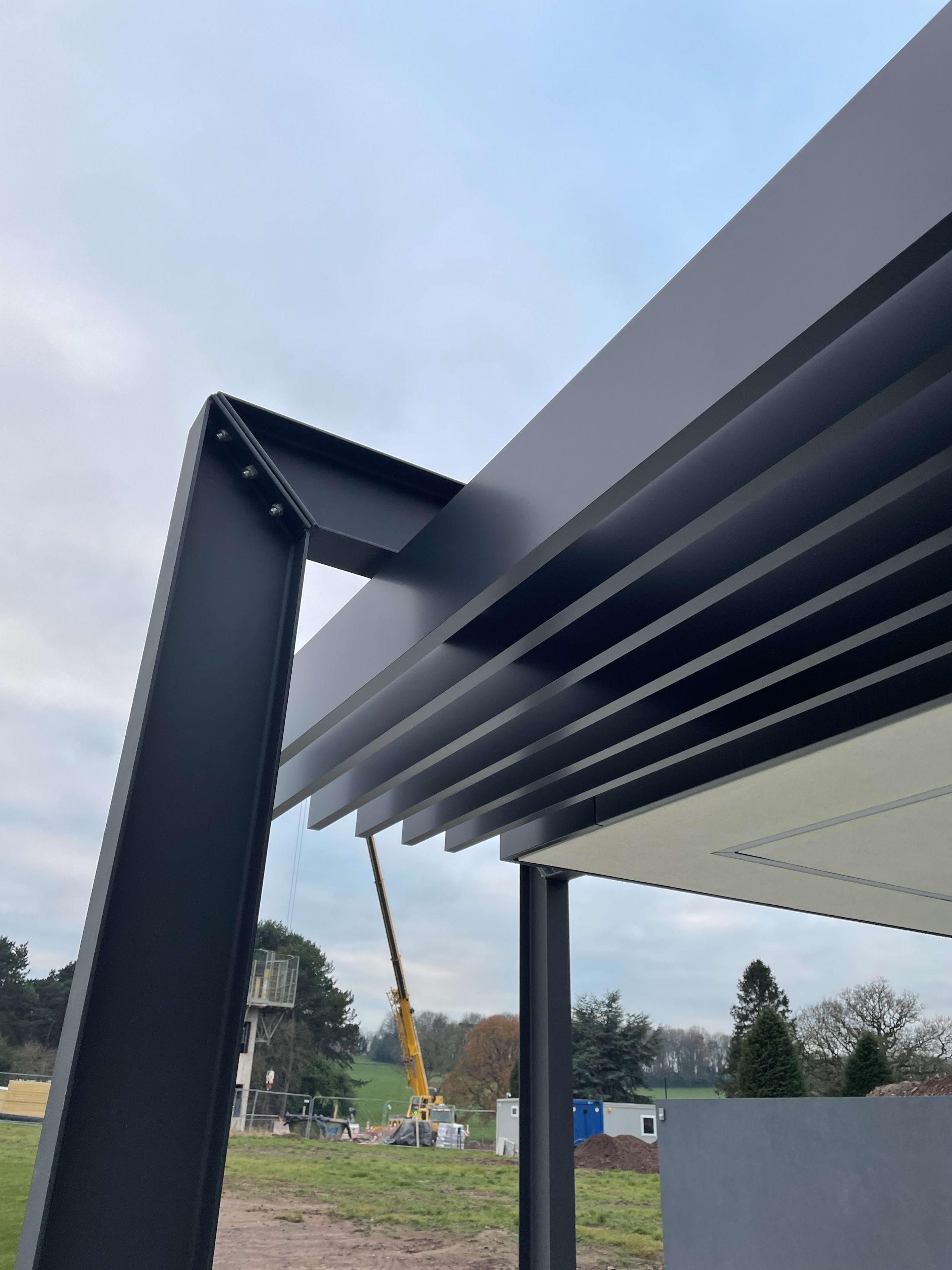 Modern anthracite aluminum pergola with louvered roof panels and angular support posts at construction site
